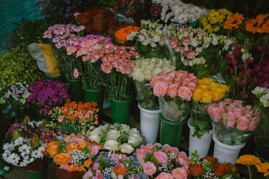 A vibrant display of fresh flowers arranged in bouquet and individual stems in various containers, including white and green pots. The flowers include peach and pink roses, white roses, white daisies
