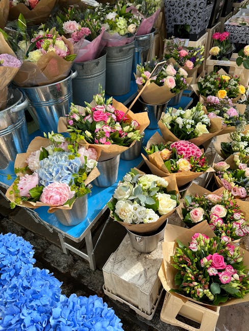 A vibrant display of fresh flower bouquets arranged in metal and glass containers, featuring a variety of pastel-colored roses, pink carnations, white lisianthus, blue hydrangeas, and green foliage, s